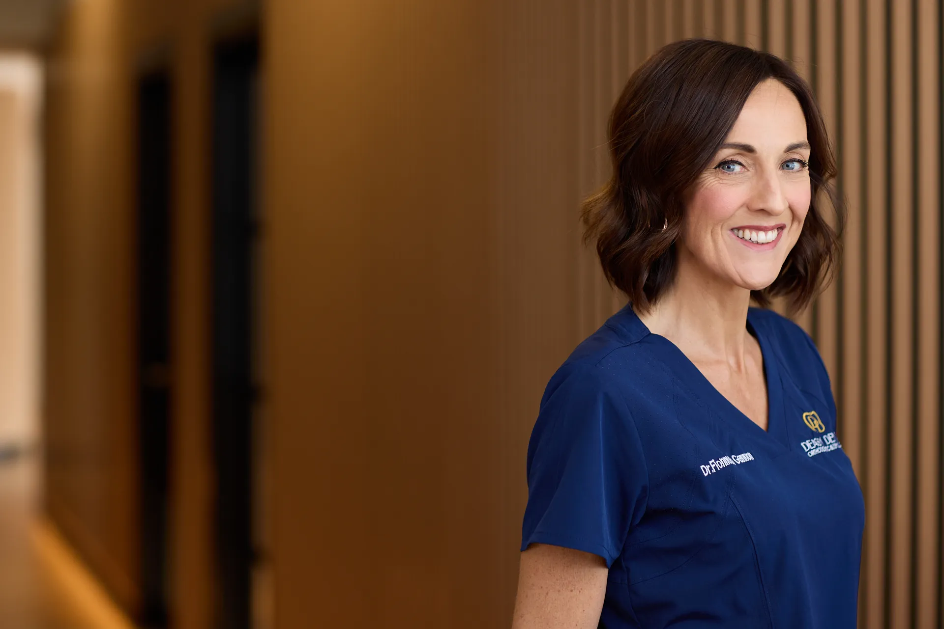 business headshot for dental practice. Woman wears branded scrubs and smiles at the camera in her place of work